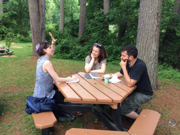 Three people talking, sitting around a picnic table, surrounded by trees